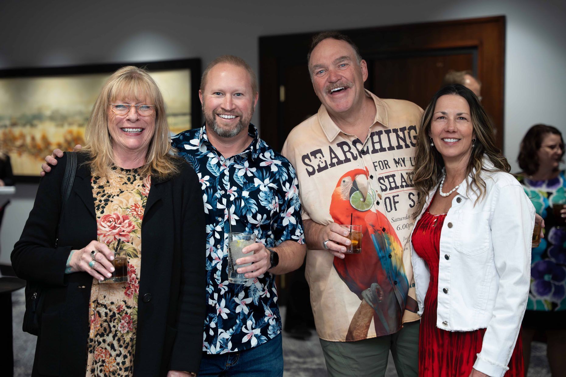 Four smiling people at an event. Two women, two men. Drinks in hand.  One man wears a parrot-themed shirt.