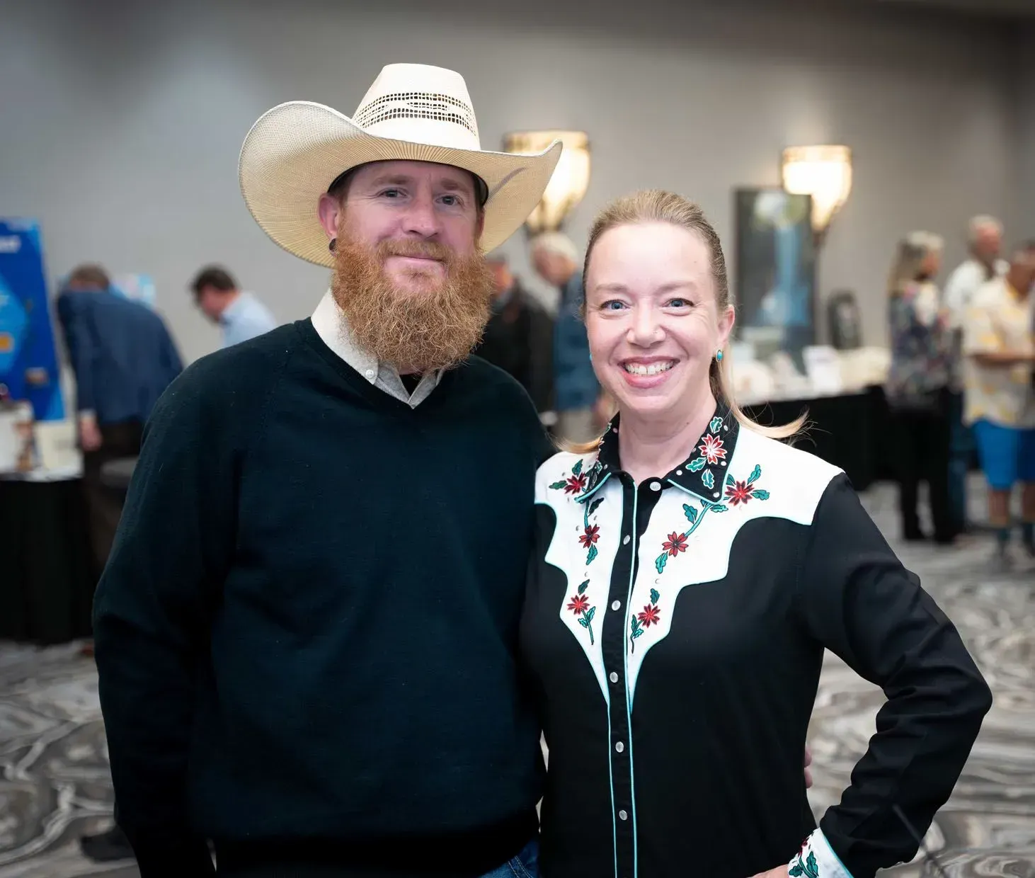 Man with a beard in cowboy hat and woman in Western shirt pose indoors, smiling.
