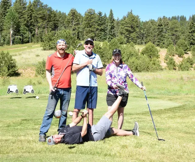 Group of four golfers posing on a green: three standing with clubs, one lying on the ground, laughing.