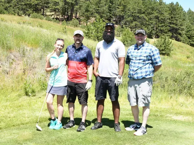 Four people posing on a golf course. Green grass, blue sky. Smiling, wearing golf attire.