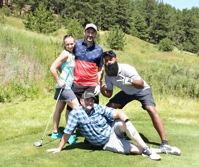 Group of four people posing on a golf course. They wear golf attire, with varied expressions and actions. Green grass and trees in background.