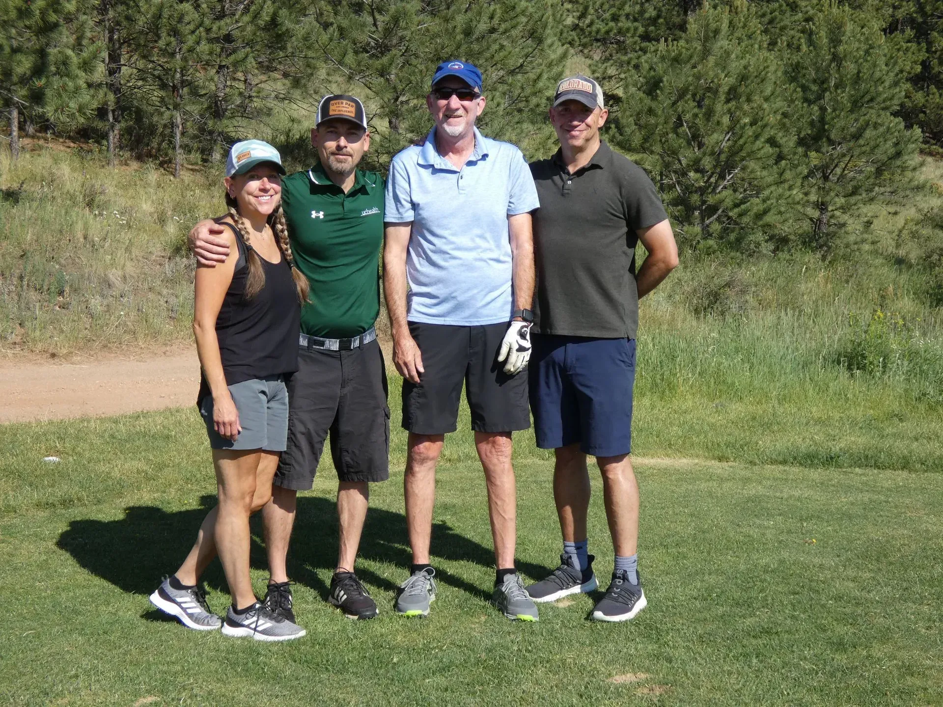 Four golfers on a green field, smiling, and posing for a photo in the sun, near trees.
