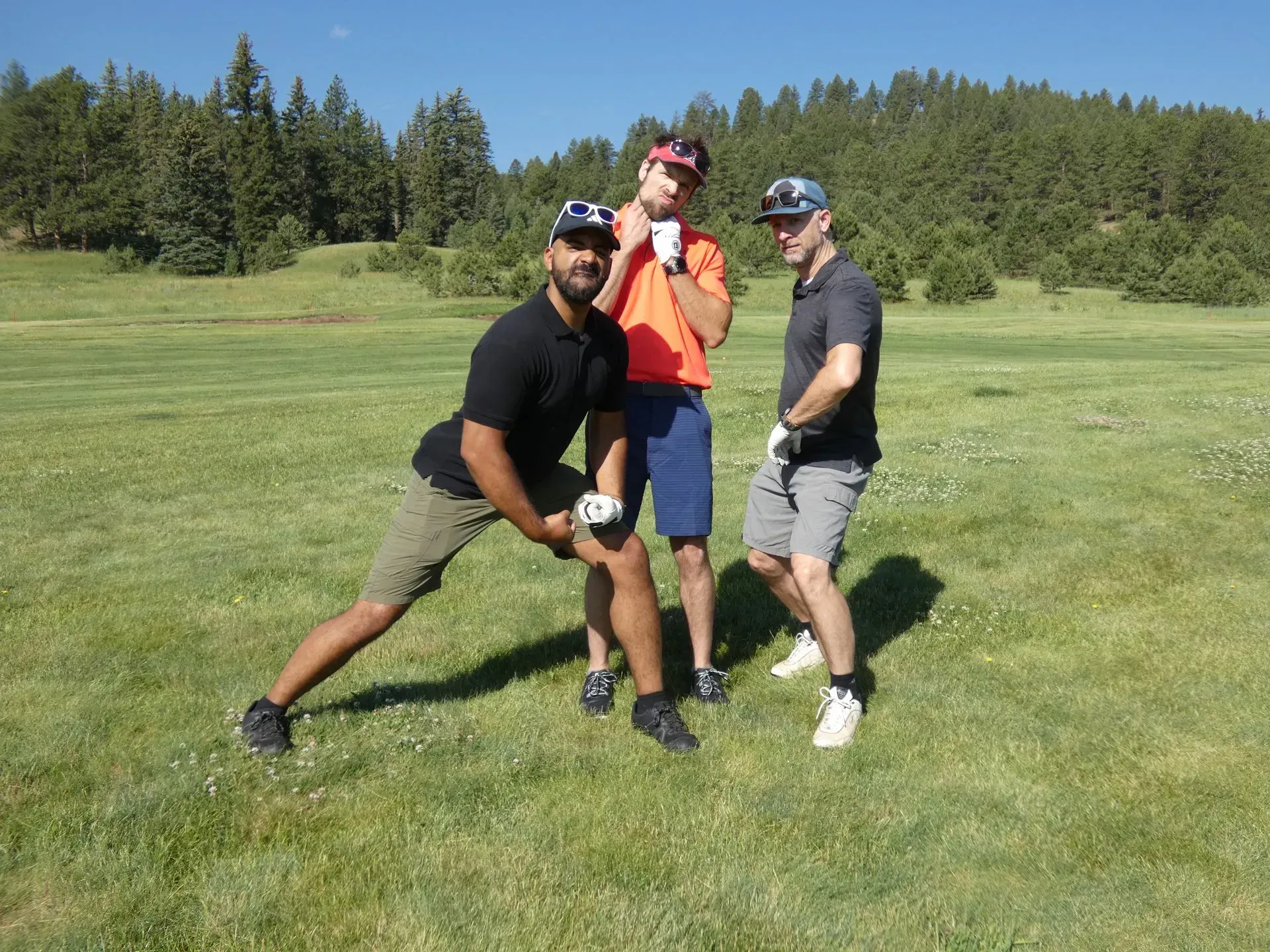 Three men posing on a golf course, the one on the left is stretching. Sunny day, green grass, trees in background.
