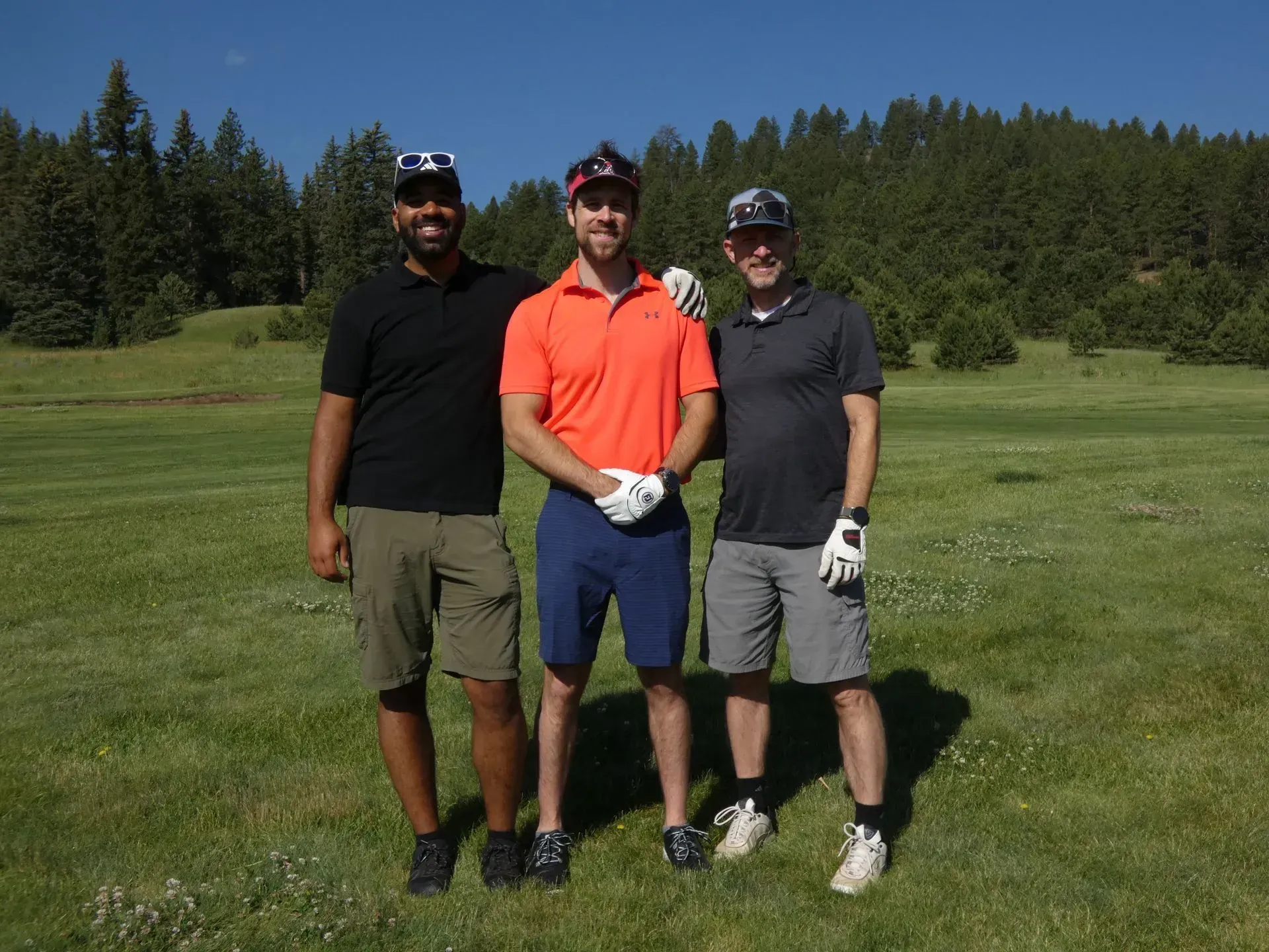 Three men on a golf course pose for a photo. They wear golf attire, and the day is sunny.
