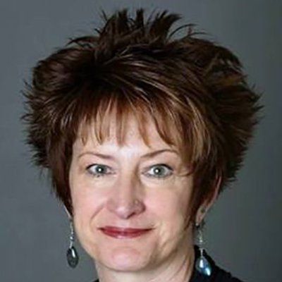 Woman with spiky brown hair, smiling, wearing earrings, against a gray background.