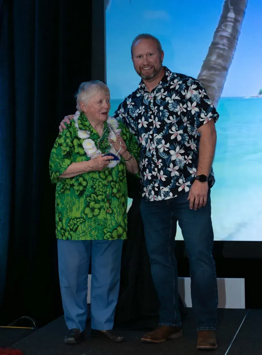 Woman in floral shirt and lei, receiving award from man in floral shirt on stage with tropical backdrop.