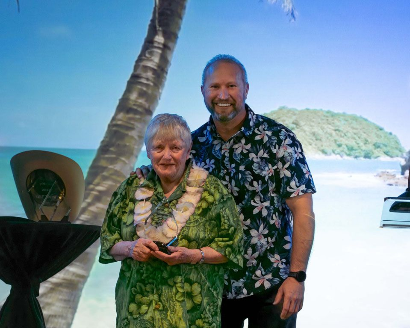 Woman in turquoise jewelry and patterned shirt receives an award from a man in a blazer.