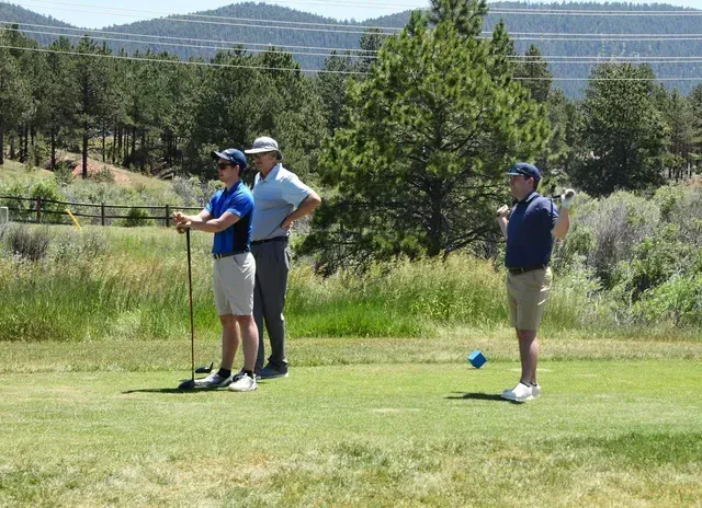 Three golfers on a green, one preparing to swing, with an instructor nearby. Sunny day, surrounded by trees.