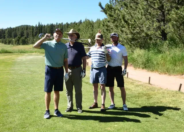 Four men on a golf course: one holding up a club, smiling, in a green shirt and shorts, and another in a cowboy hat.
