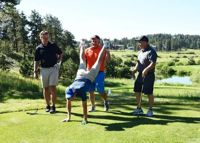 Man doing a handstand on a golf course, watched by three other men in bright daylight.