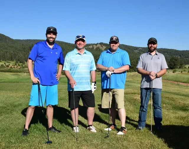 Four men on a golf course, holding clubs and posing in bright sunlight. Blue sky and green field visible.