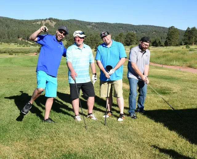 Four men golfing on a sunny course: Blue shirts, shorts, and hats. One man poses, others ready to play.