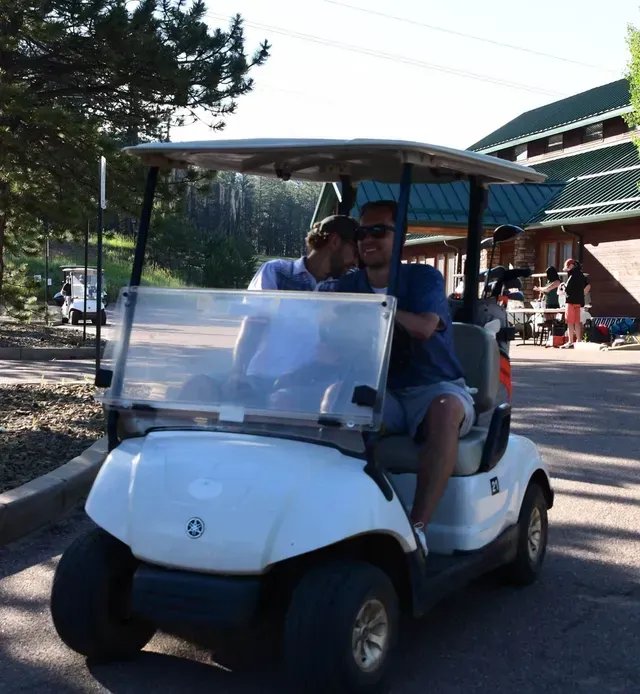 Two men in a white golf cart, one driving, on a golf course with trees and a building in the background.