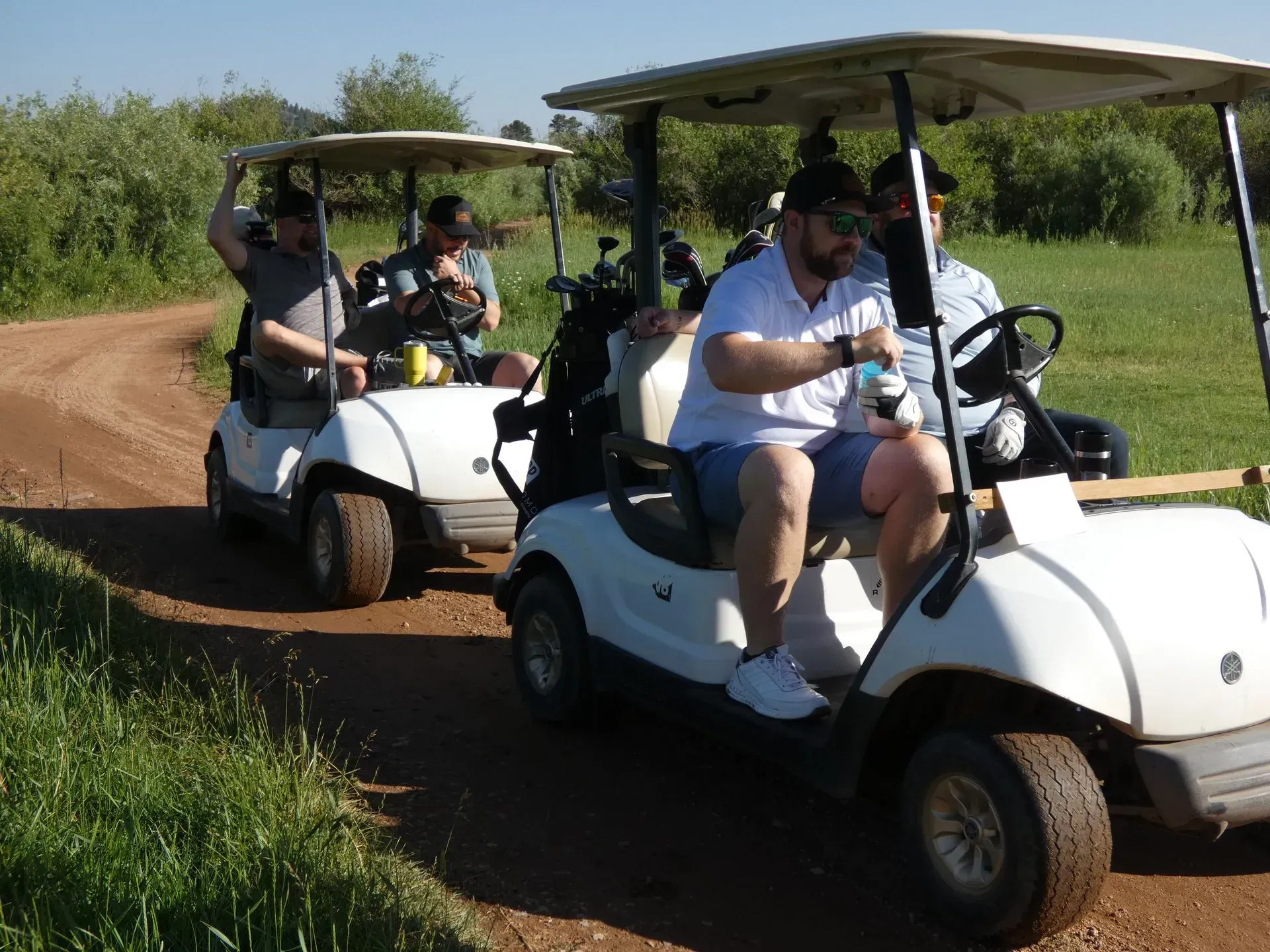 Four men in golf carts on a dirt path. The men appear to be playing golf on a sunny day.