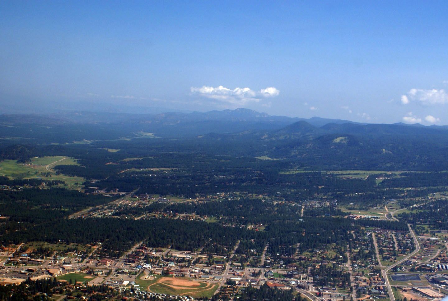 Aerial view of a town nestled in a valley, surrounded by green trees and mountains under a blue sky.