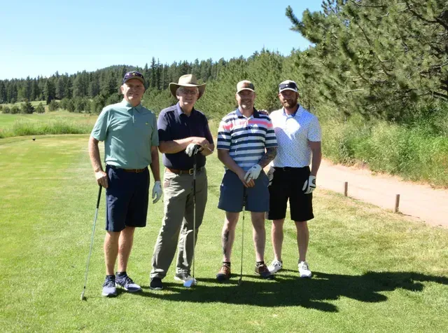 Four men standing on a golf course. They wear golf attire, smiling, and holding clubs on a sunny day.