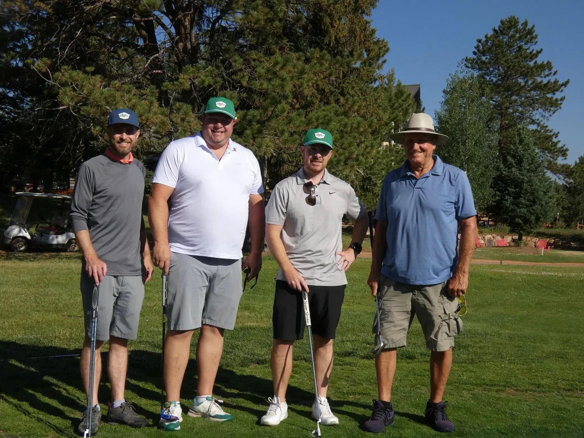 Four men on a golf course, wearing casual attire. They are holding golf clubs, smiling in the sun.