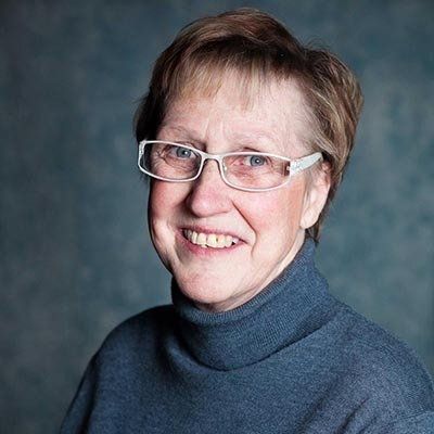 Smiling woman with glasses in a blue turtleneck, short blonde hair, against a blurred blue background.