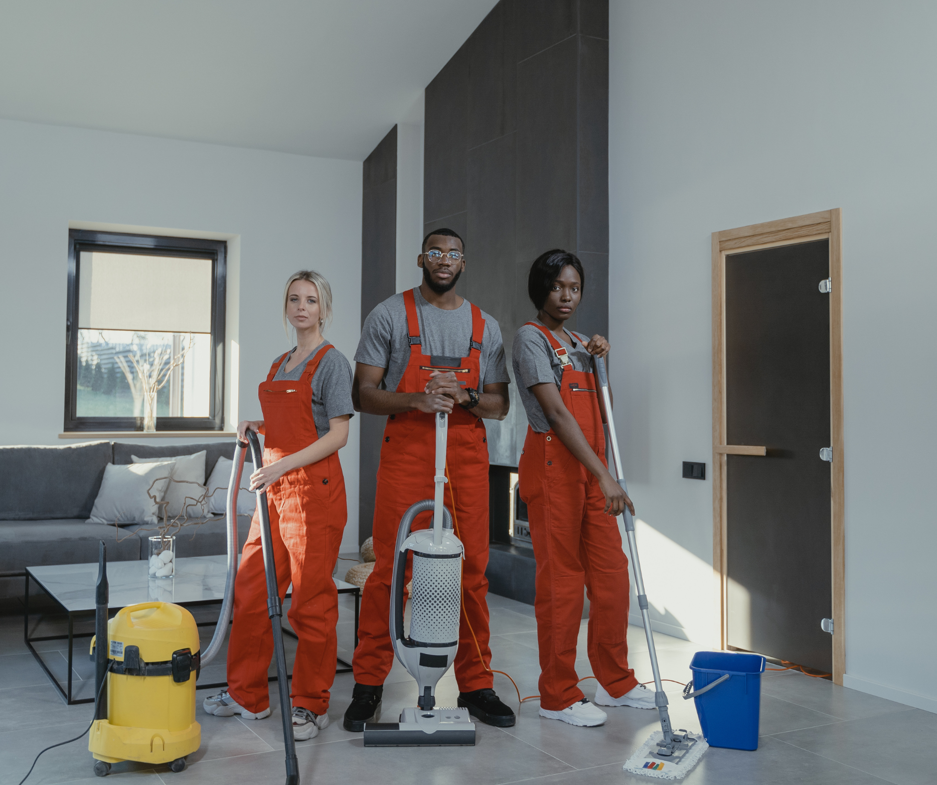 Three people in red jumpsuits pose with cleaning supplies in a modern home.