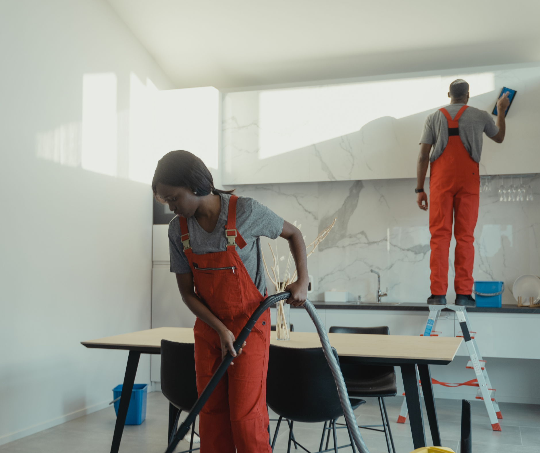 Two people in orange overalls clean a modern kitchen: one vacuums, the other scrubs a wall while standing on a ladder.