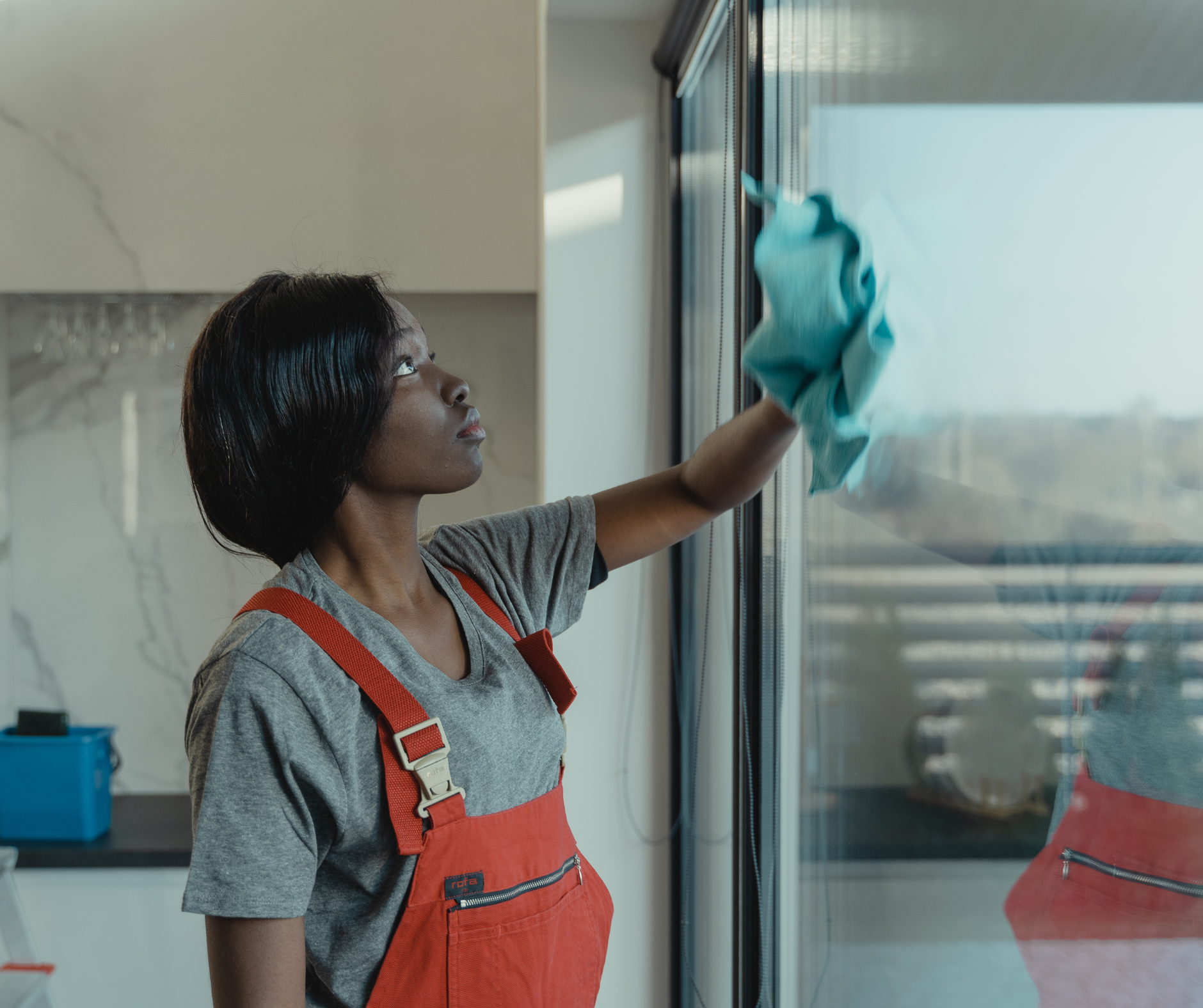 Woman in red overalls cleaning a window with a blue cloth, in a bright room.