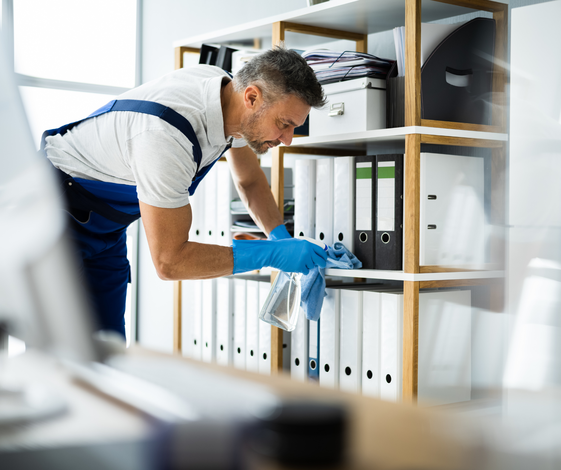 Man wearing gloves cleans office shelf with spray bottle and cloth.