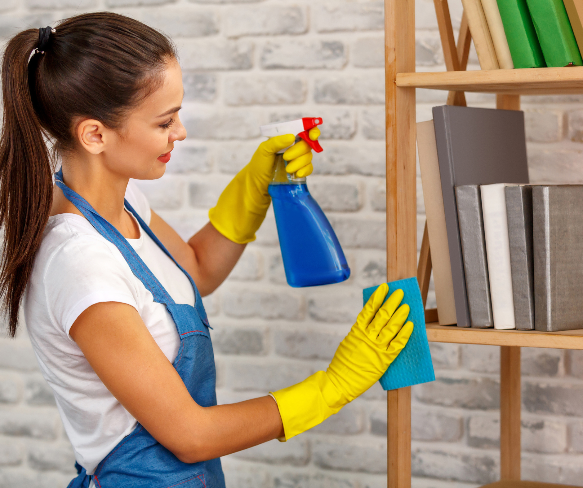 Woman in yellow gloves cleaning wooden bookshelf with spray bottle and cloth.