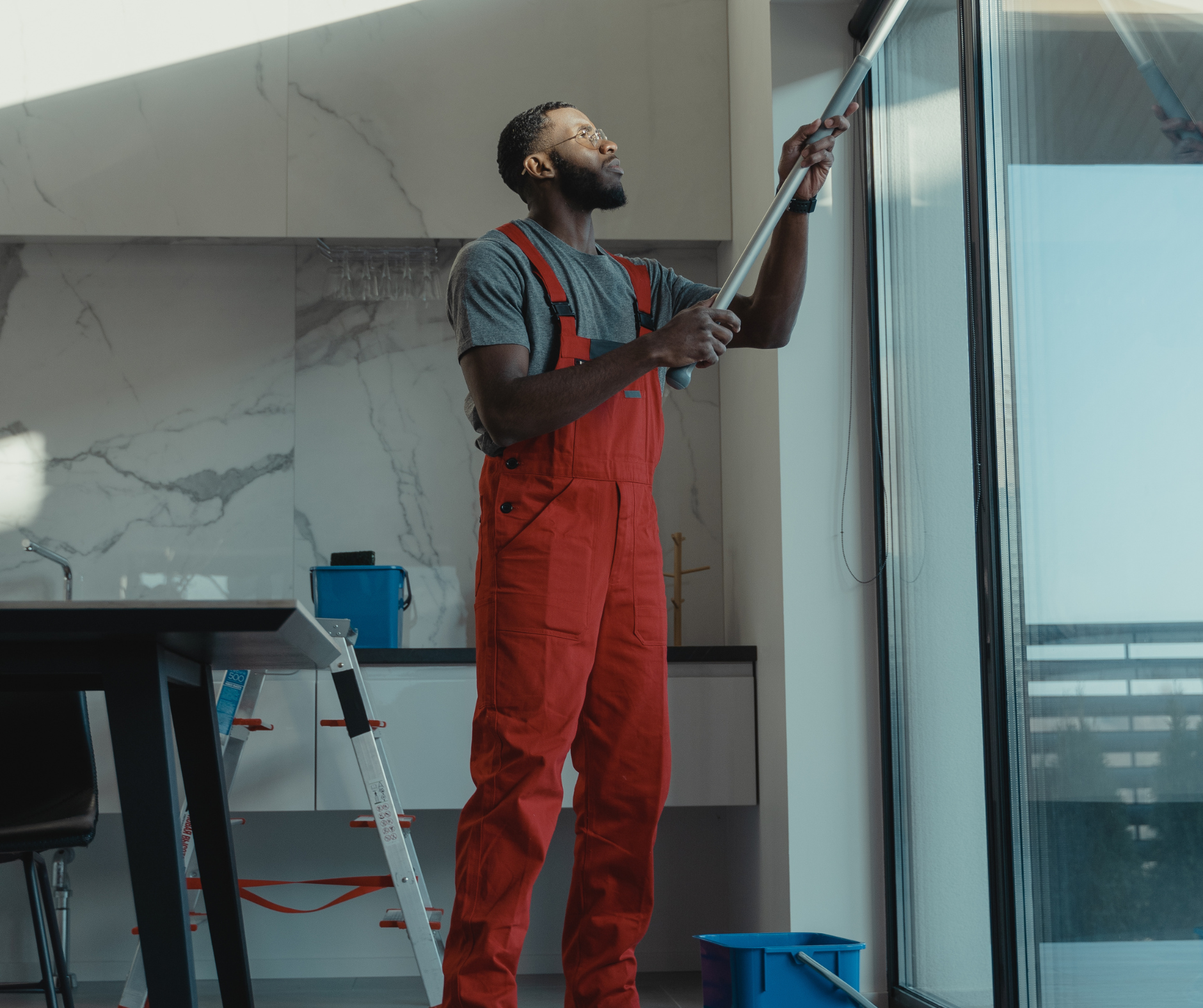 Man in red overalls cleans a tall window with a squeegee inside a modern kitchen.
