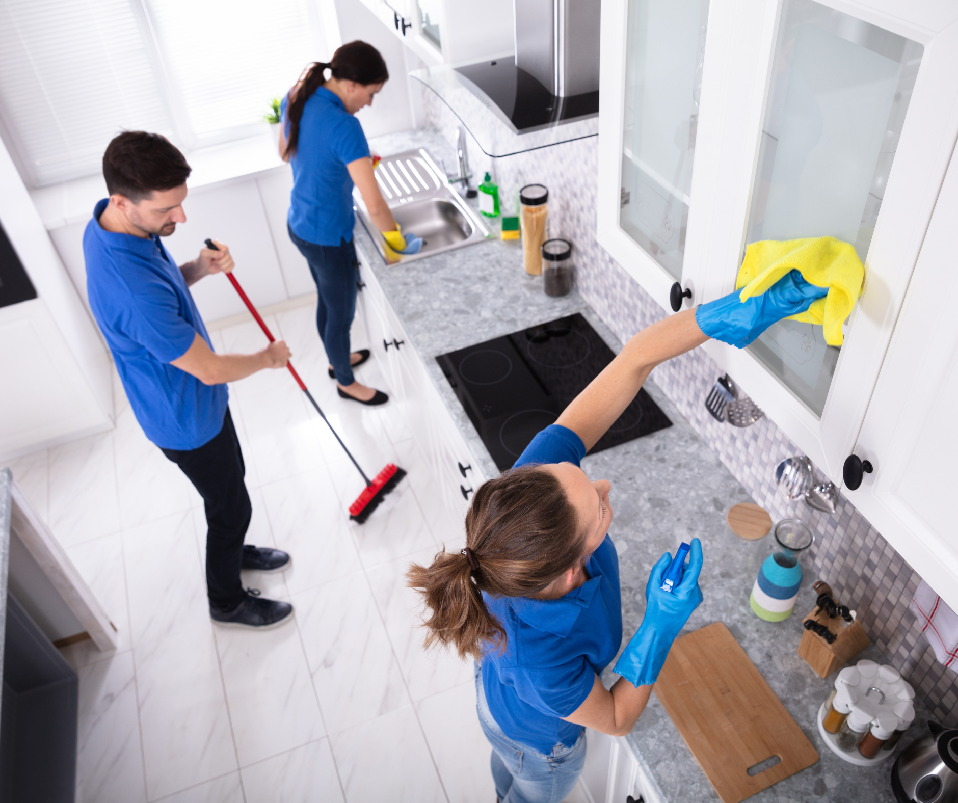 Three people cleaning a kitchen; one sweeping, one washing dishes, and one wiping cabinet doors.
