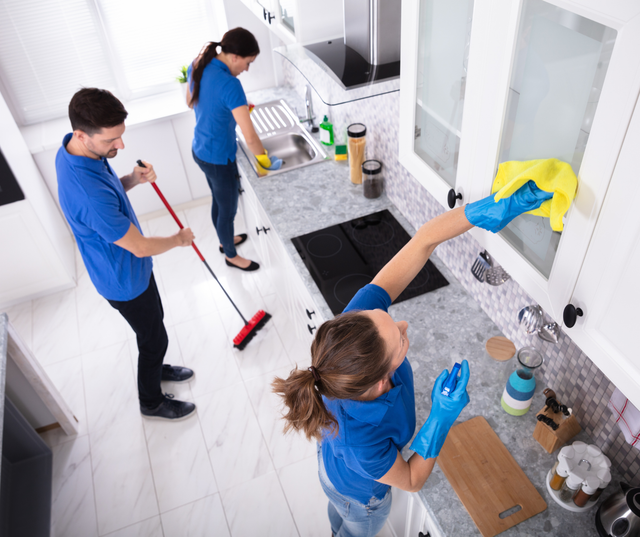 Three people cleaning a kitchen; one sweeping, one washing dishes, and one wiping cabinet doors.