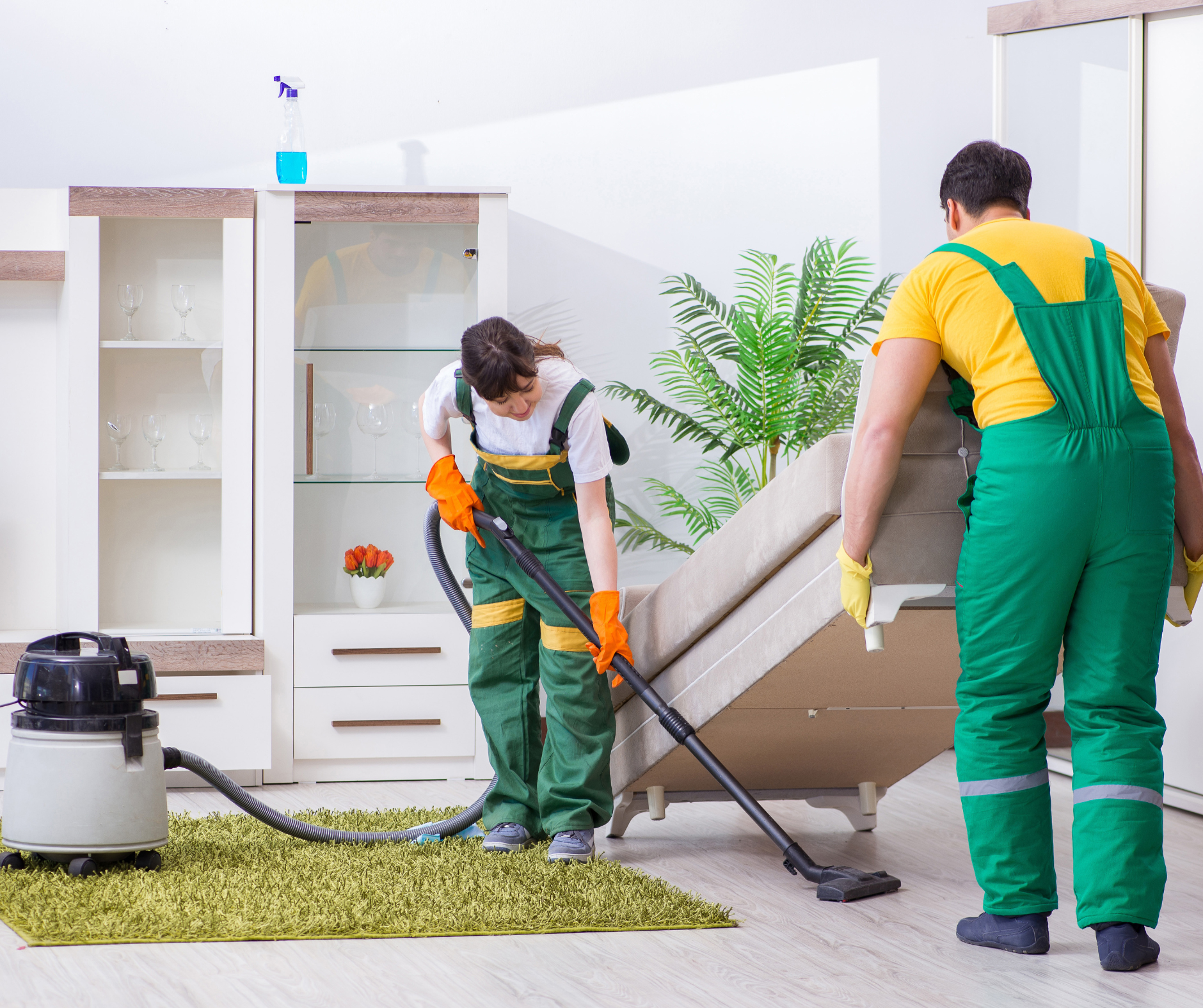 Two people cleaning a living room: one vacuuming the rug, the other lifting a sofa.
