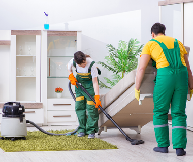 Two people cleaning a living room: one vacuuming the rug, the other lifting a sofa.