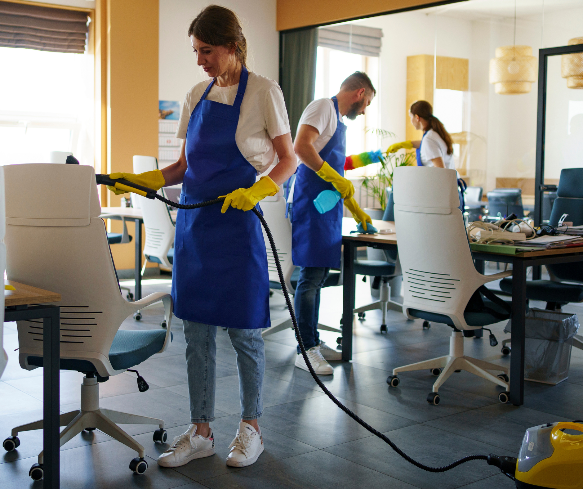 Three people cleaning an office, wearing aprons and gloves, with cleaning equipment.
