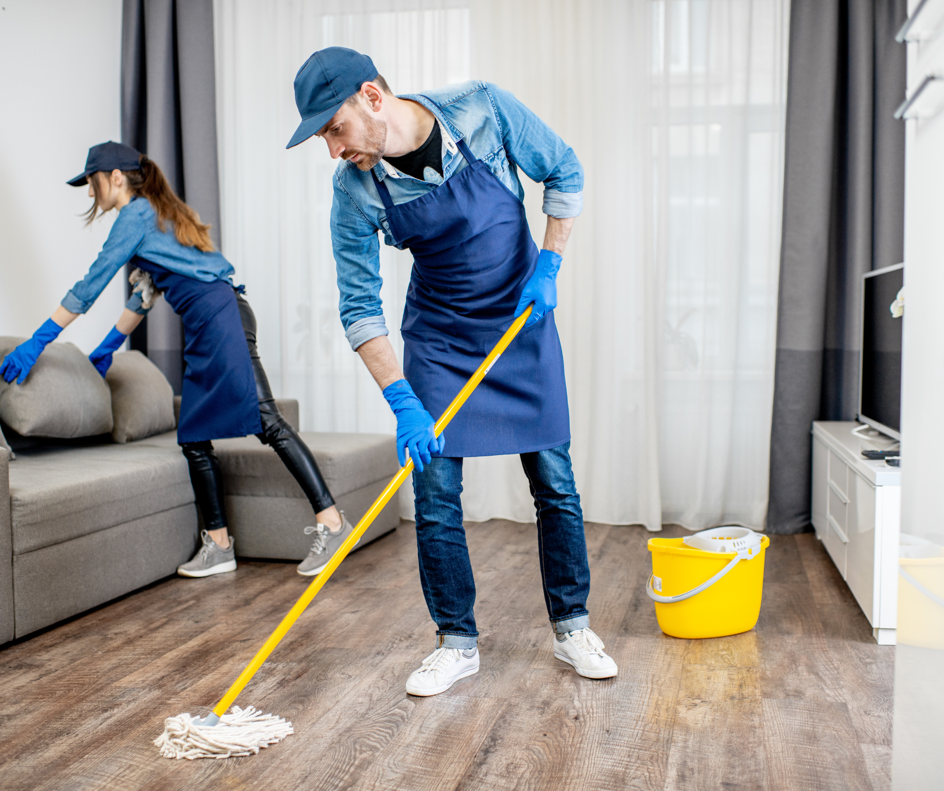 Two people in cleaning uniforms mop a room; a yellow bucket sits nearby.