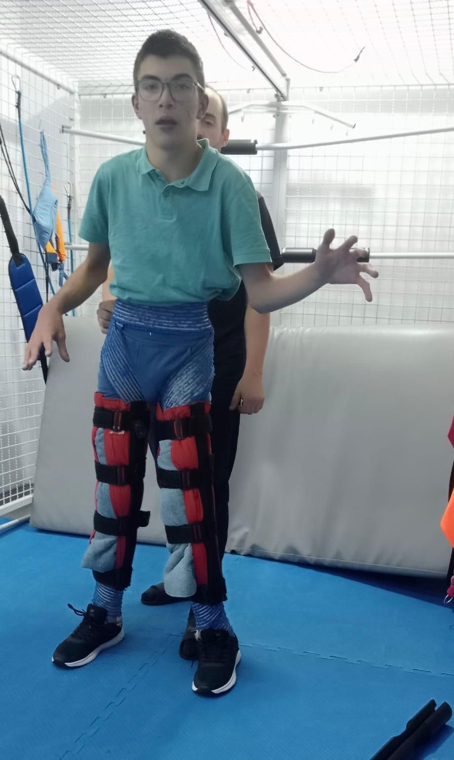 Menino com aparelhos ortopédicos nas pernas, amparado por um auxiliar, está em uma sala de terapia, gesticulando com a mão.