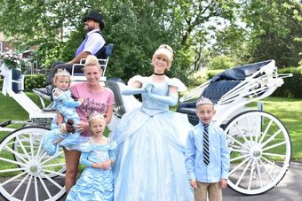 Woman in a light blue gown with children in front of a white horse-drawn carriage outdoors