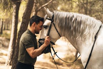 Man gently touching noses with a white horse in a wooded setting