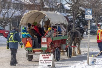 Horse-drawn wagon with passengers on a snowy street, guided by workers in safety vests.