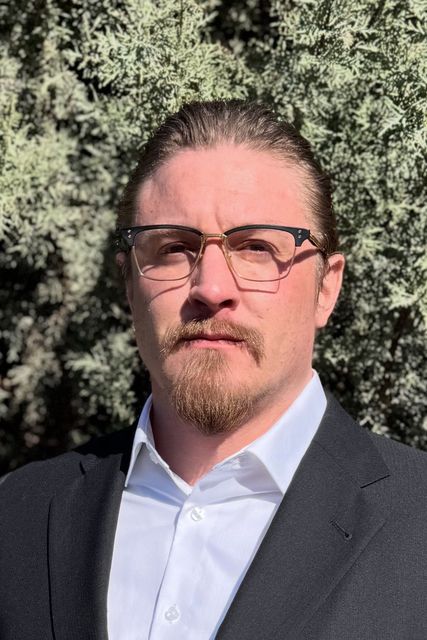 A man wearing glasses and a suit is standing in front of a bush.