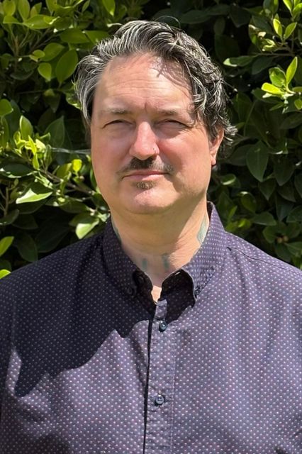 A man with a mustache and tattoos is wearing a white shirt.