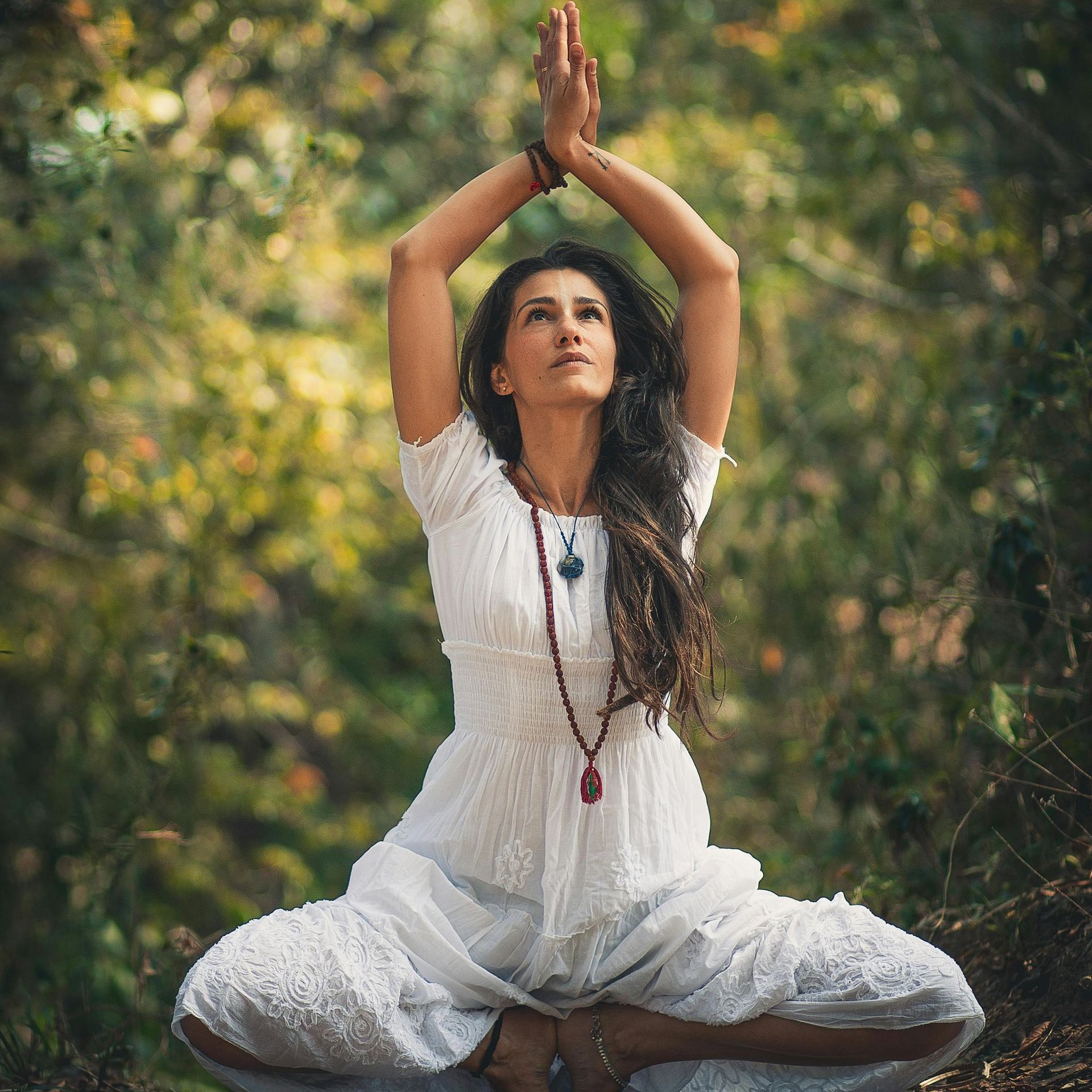 A woman in a white dress is sitting in a lotus position with her hands in the air.