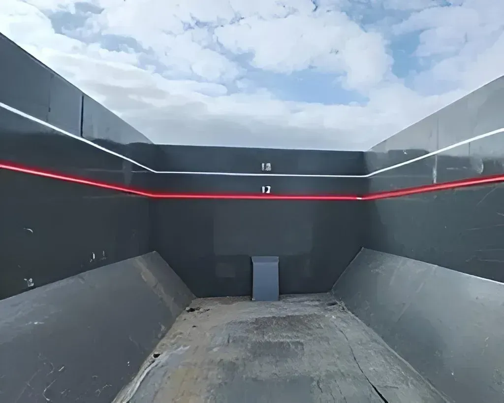 Empty dark grey interior of a large metal dump truck bed with white horizontal capacity markers and a red top edge.