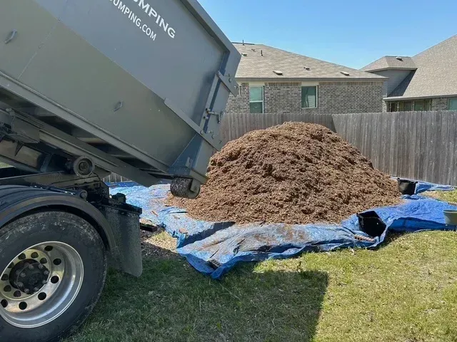 A dump truck is unloading a large pile of brown mulch onto a blue tarp in a grassy residential backyard.
