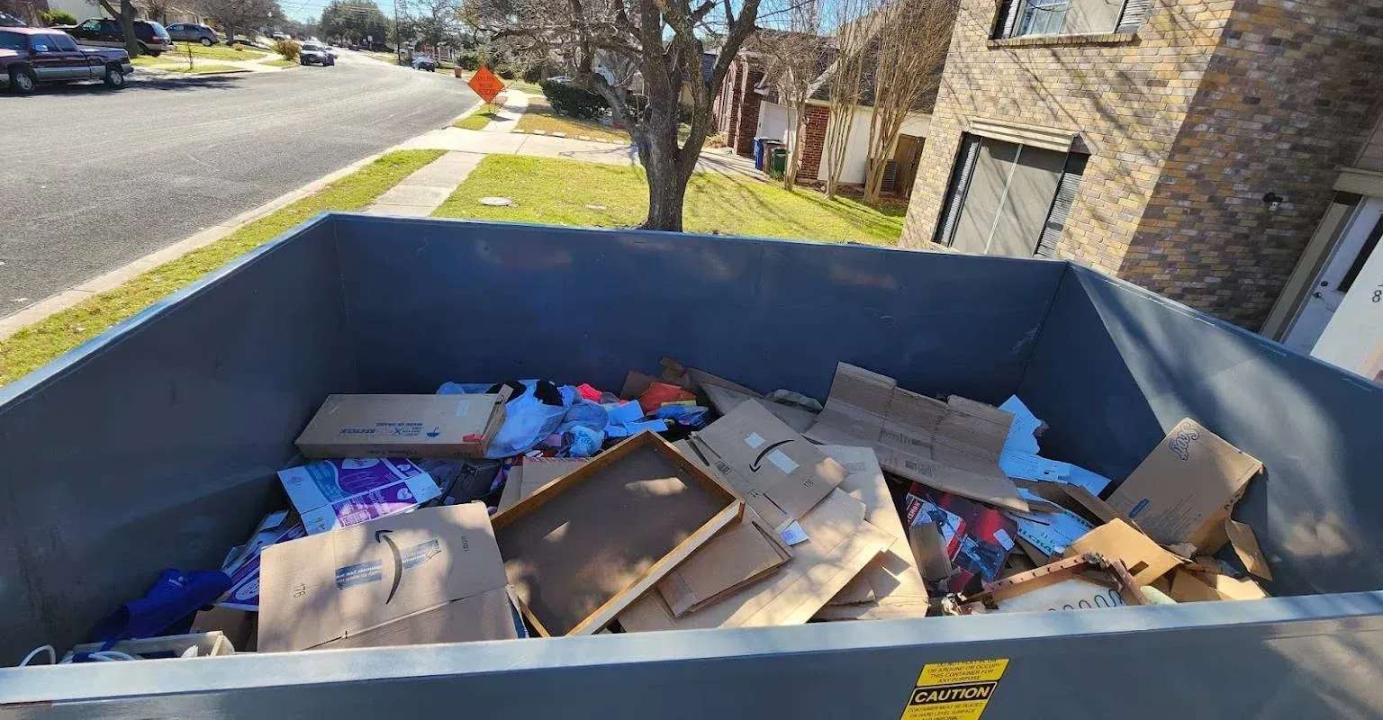 A gray dumpster sits on a residential street filled with cardboard boxes, paper, and various discarded items.