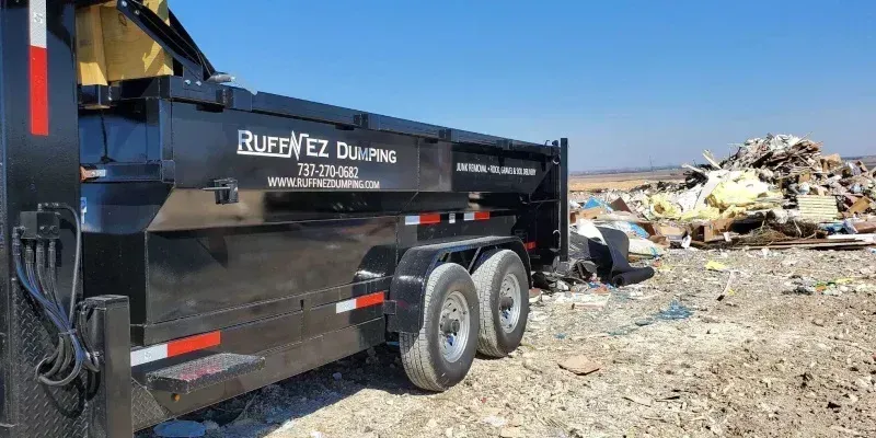 A black dump trailer parked on a dirt lot next to a large pile of debris under a clear blue sky.