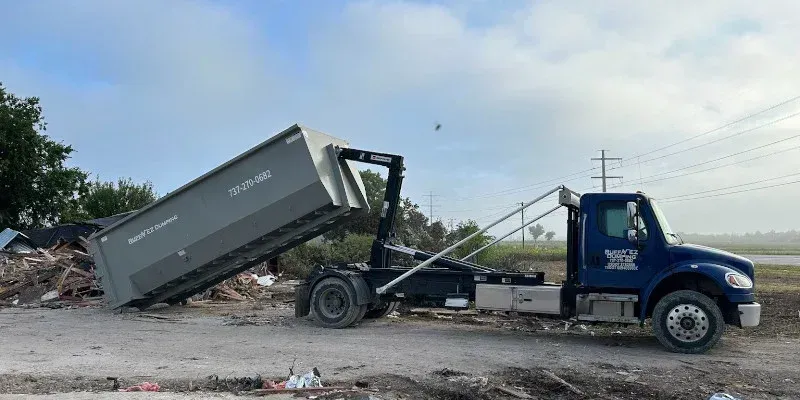 A blue roll-off dump truck in an outdoor setting, actively lowering a large grey metal dumpster onto a gravel lot.