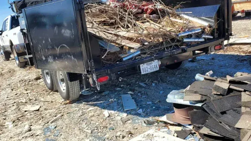 A black dump trailer filled with debris and tree branches sits on a dirt lot, with a pile of shingles in the foreground.