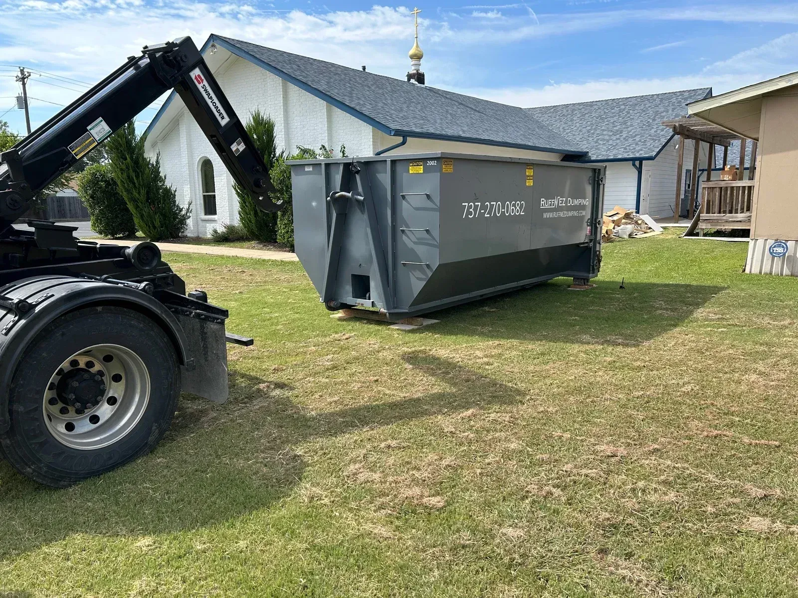 A roll-off truck positioning a large grey dumpster on a grass lawn in front of a white building under a blue sky.