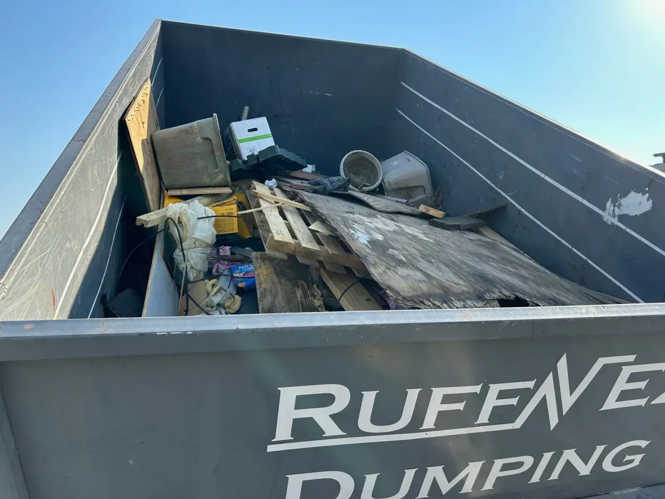 A grey RUFF-N-EZ dumpster filled with various pieces of construction debris, wood planks, and household trash.