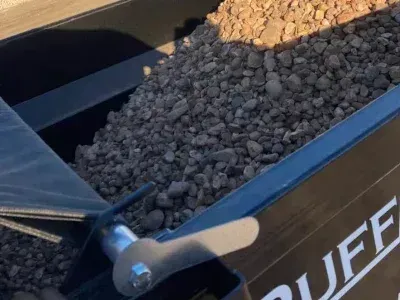 A close-up of the metal side of a black Ruffneck dump trailer filled with gray gravel, showing a silver latch mechanism.